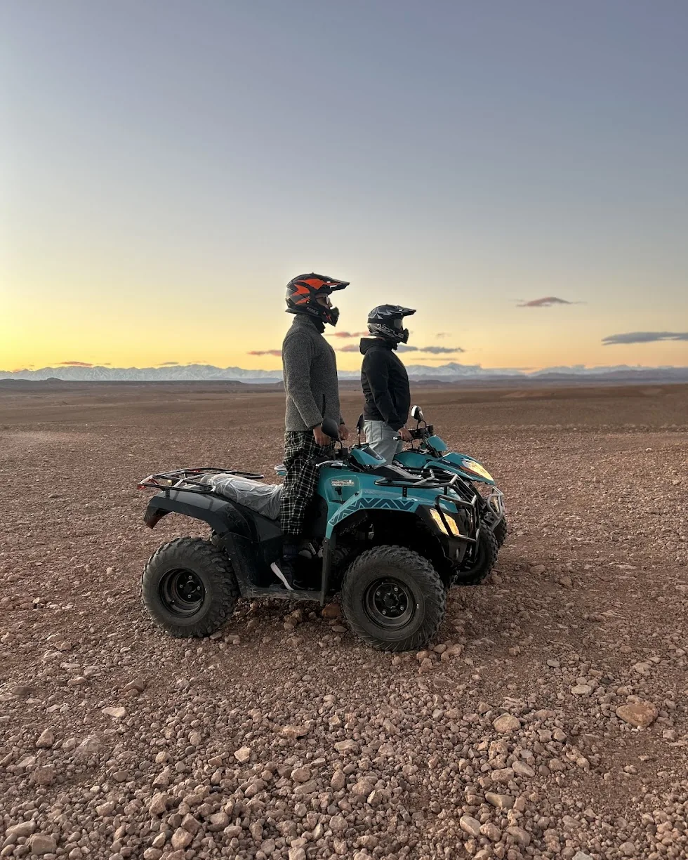 Two helmeted riders on a turquoise quad at sunset